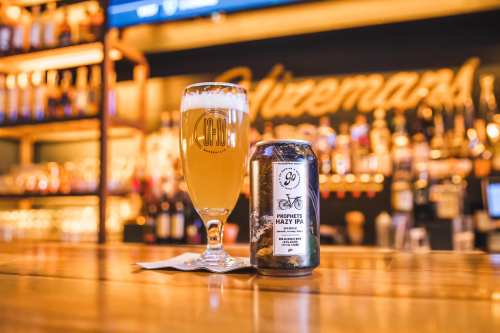 A glass of hazy IPA beer next to a can on a wooden bar, with a blurred background of bottles and warm lighting.