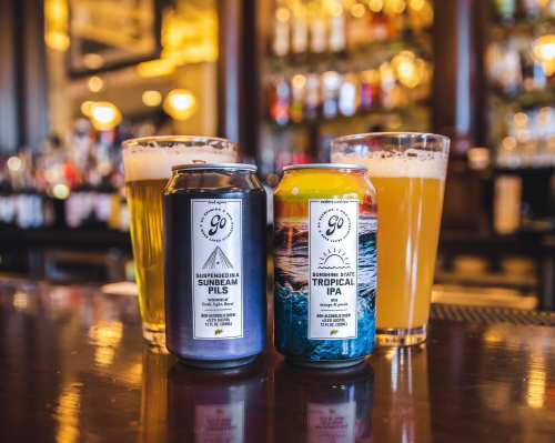 Two cans of craft beer on a bar, with glasses of beer in the background, showcasing a lively pub atmosphere.