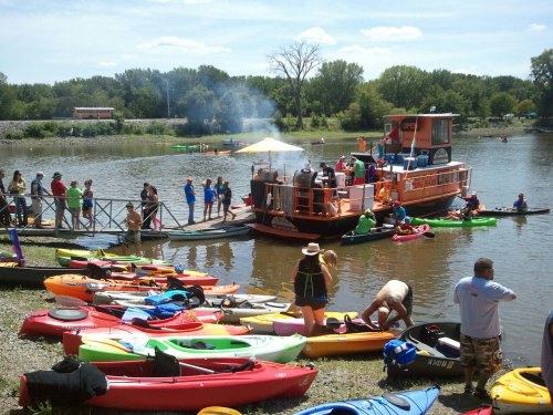 A busy river scene with colorful kayaks, people on the shore, and a boat with smoke rising from a grill.