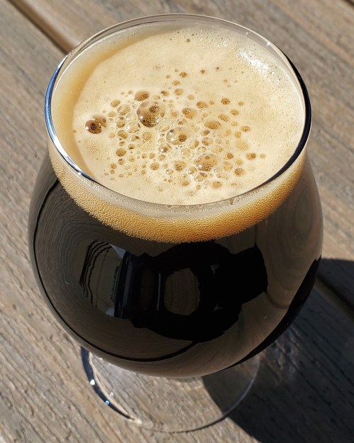 A close-up of a dark beer in a curved glass, featuring a creamy, frothy head on top, set on a wooden surface.