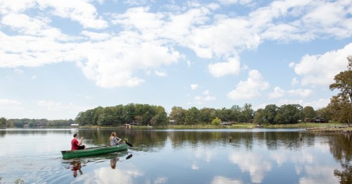Two people paddle a green canoe on a calm lake under a blue sky with fluffy clouds, surrounded by trees.
