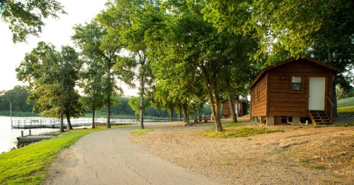 A peaceful lakeside scene with wooden cabins, a gravel path, and lush green trees under a clear sky.