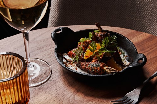 A cast iron dish with grilled vegetables garnished with herbs, alongside a glass of white wine and a decorative candle holder.