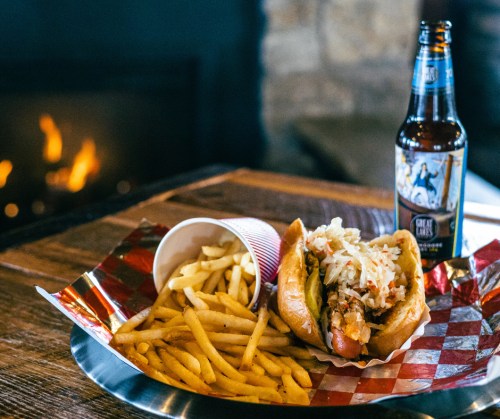 A hot dog topped with coleslaw, served with fries and a bottle of soda, on a wooden table near a fireplace.