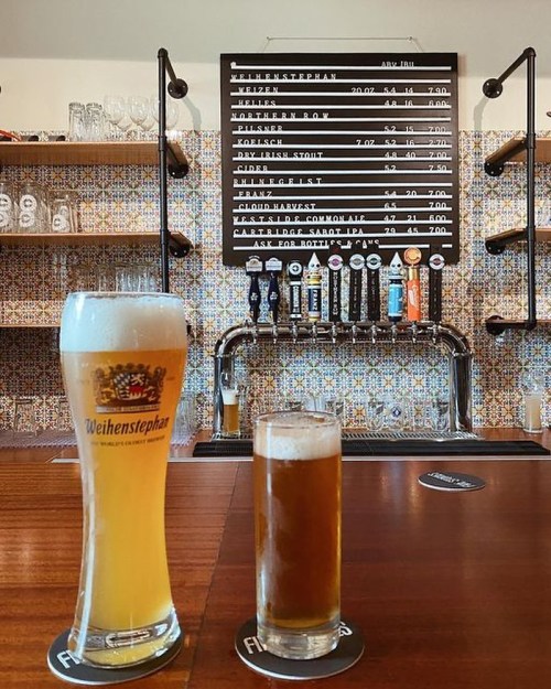 Two glasses of beer on a wooden bar, with a menu of drinks displayed on a wall behind them.