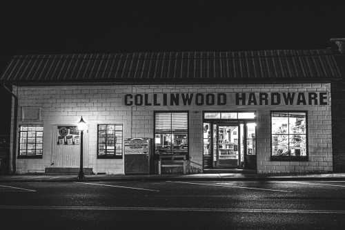 A black and white photo of Collinwood Hardware store at night, illuminated with warm light from the windows.