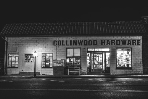A black and white photo of Collinwood Hardware store at night, illuminated with warm light from the windows.
