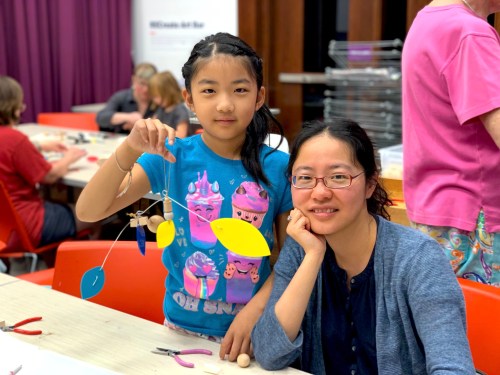 A young girl holds a colorful mobile while sitting next to a woman, both smiling at a crafting workshop.