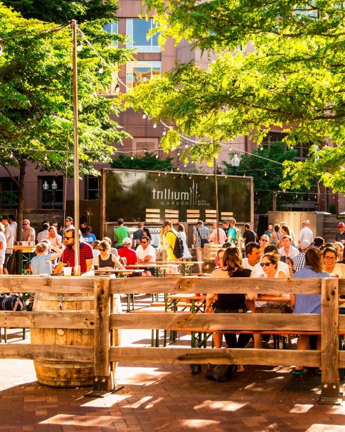 A lively outdoor garden with people dining and socializing under trees, featuring wooden tables and a vibrant atmosphere.