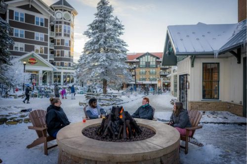 A group of people sitting around a fire pit in a snowy resort area, with buildings and trees in the background.