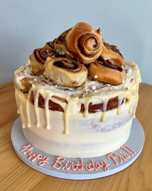 A birthday cake topped with cinnamon rolls and drizzled with icing, with "Happy Birthday Pii!!" written on the base.