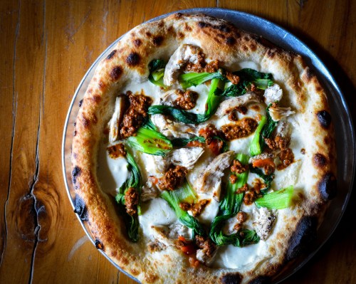 A freshly baked pizza topped with chicken, greens, and a savory sauce on a golden crust, served on a metal tray.