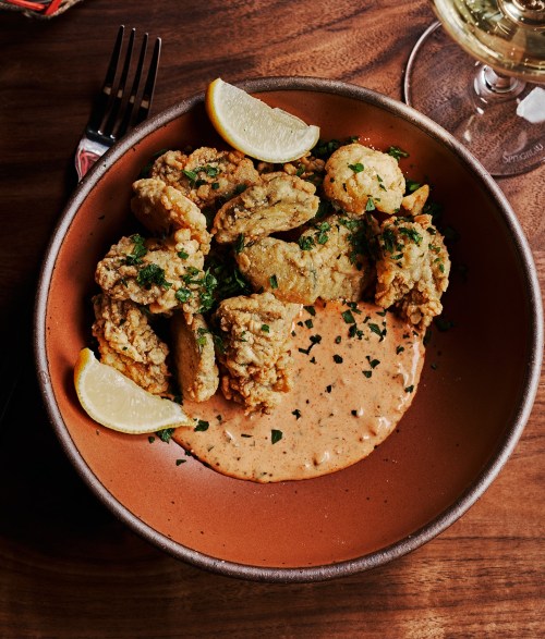 A bowl of fried chicken pieces with a creamy sauce, garnished with parsley and lemon wedges, on a wooden table.