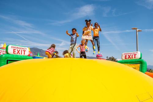 Children joyfully jumping on a large yellow inflatable, with emergency exit signs visible in a sunny outdoor setting.