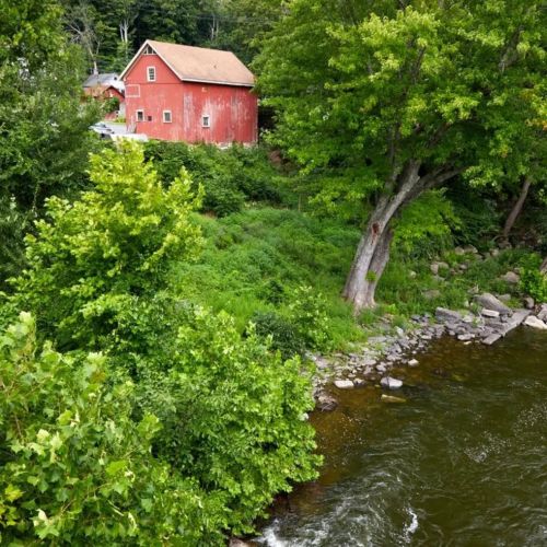 A red barn beside a lush green landscape and a flowing river, surrounded by trees and rocky shoreline.