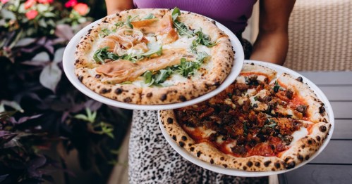 A person holds two pizzas: one topped with prosciutto and arugula, the other with sausage and greens, against a floral backdrop.