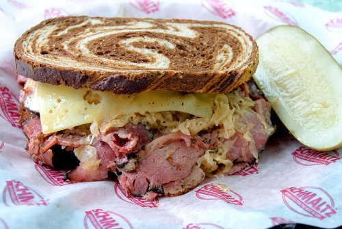 A hearty sandwich with layers of pastrami, sauerkraut, and cheese on marbled rye, served with a pickle on the side.