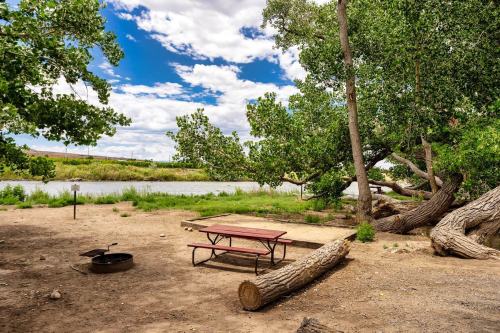A serene riverside campsite with a picnic table, fire pit, and lush trees under a partly cloudy sky.