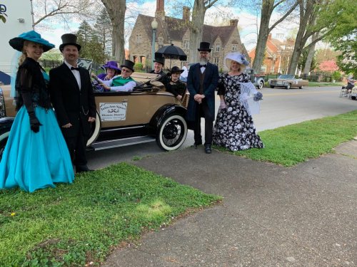 A group in vintage attire poses by a classic car on a tree-lined street, showcasing a festive, historical theme.