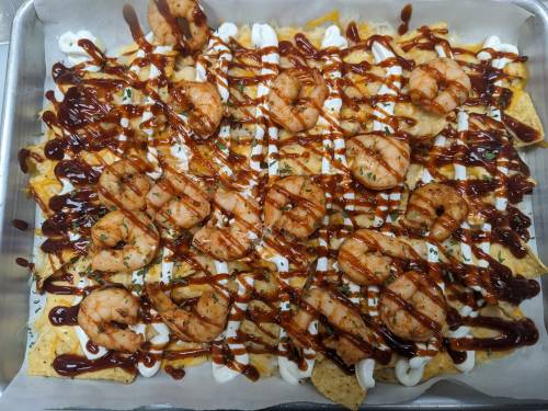 A tray of nachos topped with grilled shrimp, drizzled with barbecue sauce and sour cream, garnished with herbs.