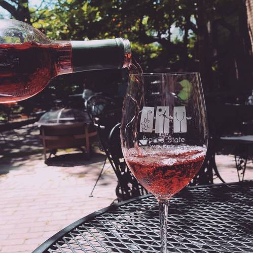 A bottle of rosé wine is being poured into a glass on a sunny outdoor patio with tables and chairs.