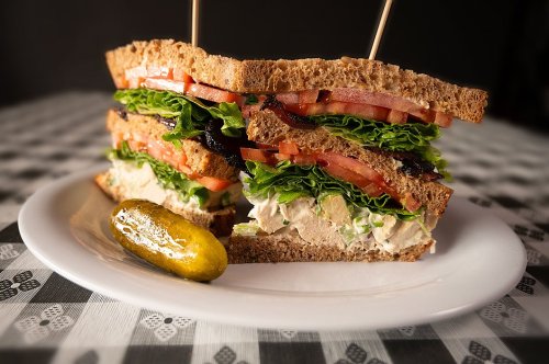 A hearty sandwich with layers of lettuce, tomato, and chicken salad on whole grain bread, served with a pickle on the side.
