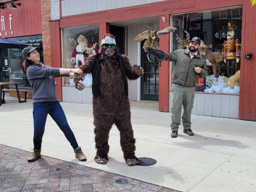 A person in a Bigfoot costume stands between two people, one playfully pulling their arm and the other holding an owl.
