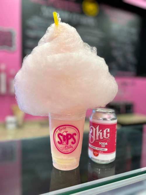 A cup of pink cotton candy topped drink next to a can of soda, set against a pink background.
