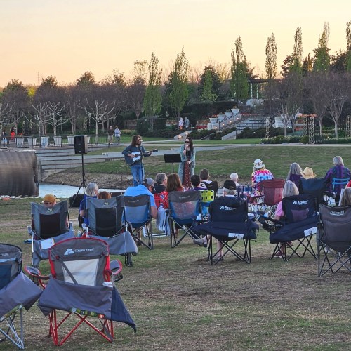 A singer and guitarist perform on stage for an audience seated in lawn chairs at an outdoor event during sunset.
