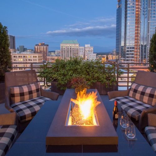 A rooftop patio with modern seating, a fire pit, and a city skyline in the background during twilight.
