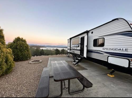 A camper parked near a scenic view, with a picnic table and trees, overlooking a lake at sunset.