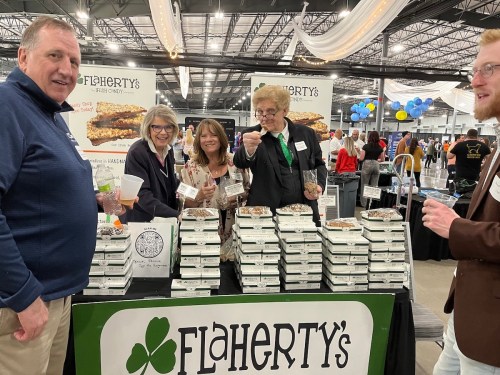 A group of five people at a food booth, smiling and holding samples, with boxes of food displayed behind them.