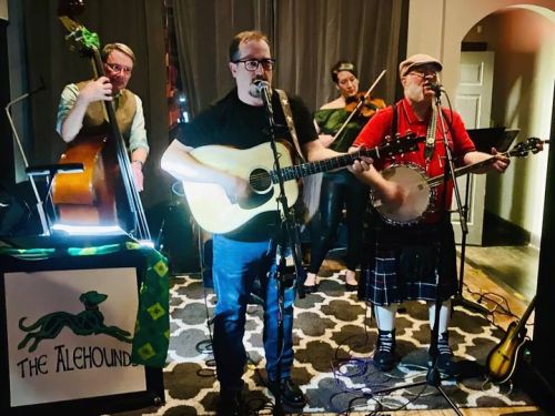A band performs on stage with guitar, banjo, and violin, featuring a man in a kilt and a backdrop with "The Alehounds."