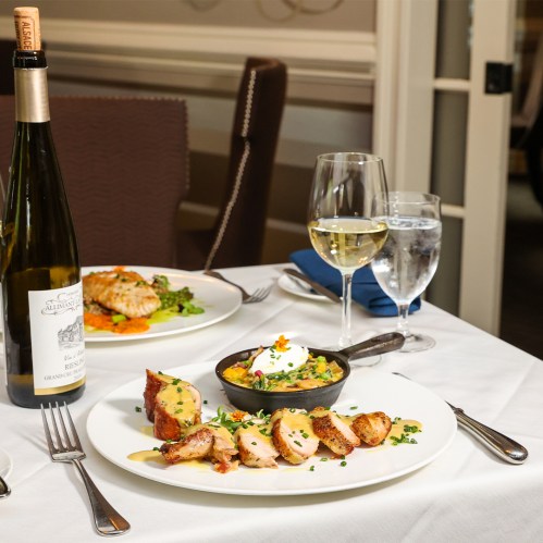 A beautifully plated meal featuring sliced chicken, a side dish in a skillet, wine, and glasses of water on a table.