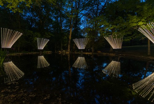 Illuminated light installations reflect in a calm pond surrounded by trees at dusk.