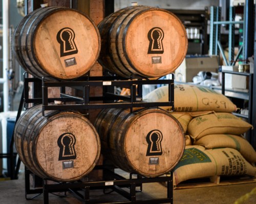 Four wooden barrels with keyhole logos stacked on a metal rack, surrounded by burlap sacks in a warehouse setting.