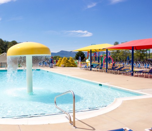 A colorful pool area featuring a water fountain, lounge chairs, and shaded umbrellas under a clear blue sky.