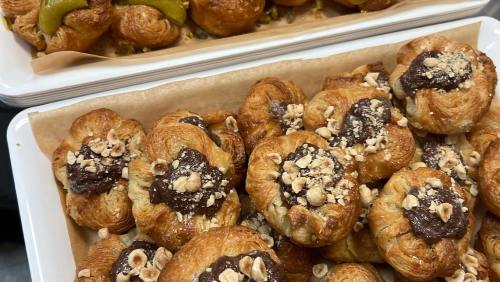 A close-up of flaky pastries topped with chocolate and crushed nuts, displayed on a tray with other baked goods in the background.