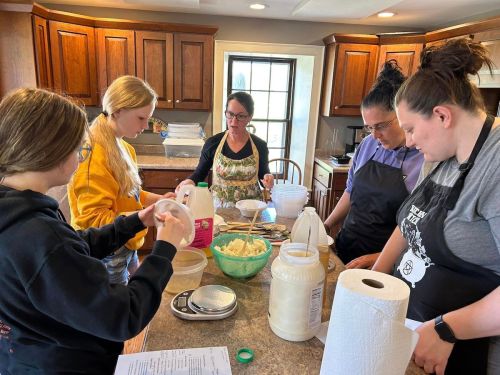 A group of five women in a kitchen preparing food, measuring ingredients, and working together at a countertop.