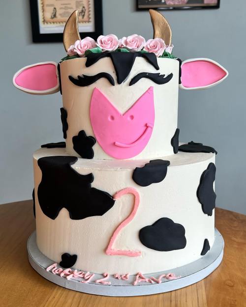 A two-tiered cow-themed birthday cake with pink accents, a floral crown, and a "2" on the front.