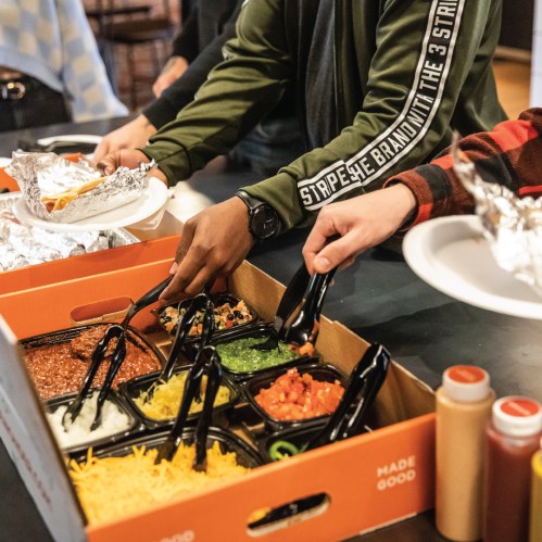 People serving food from a large box with various toppings and sauces, using tongs to fill their plates.