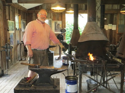 A smiling blacksmith stands in a rustic workshop, next to an anvil and a forge with a small fire burning.