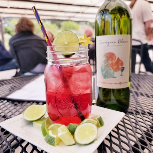 A refreshing drink in a mason jar with lime slices, a straw, and a flower, alongside a bottle of Sauvignon Blanc.