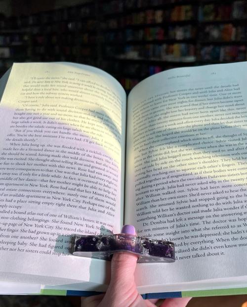 A hand holds an open book with highlighted text, illuminated by soft light, against a blurred bookshelf background.