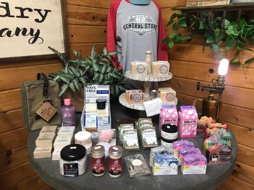 Display of various soaps and personal care products on a table, with a shirt and decorative plants in a rustic setting.