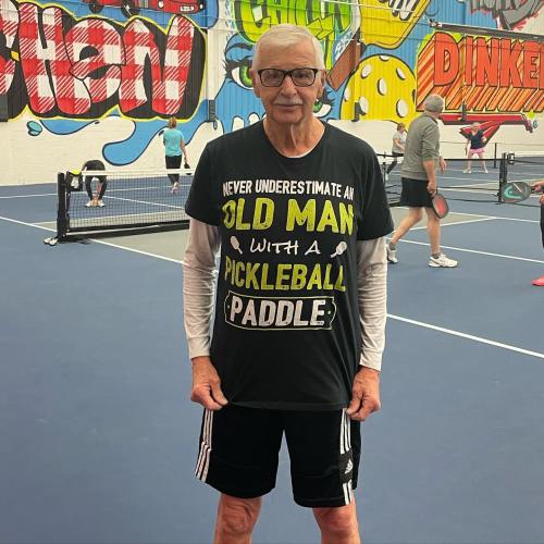An older man in a black pickleball t-shirt stands on a court, smiling with a paddle in hand.