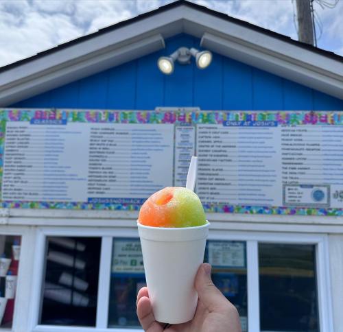 A hand holds a colorful snow cone in front of a blue building with a menu displayed on the wall.