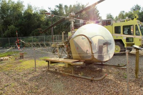 A vintage helicopter with a transparent cockpit displayed outdoors next to a fire truck in a gravel area.