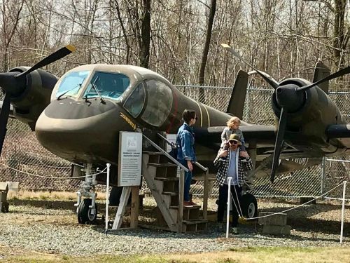 Visitors stand near a vintage military aircraft displayed outdoors, surrounded by trees and a fence.