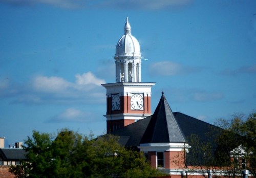 A clock tower with a white dome and a black roof, surrounded by trees under a blue sky with clouds.